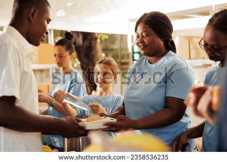 Women of different ethnicities volunteering at local food bank serving food and non-perishable items to the hungry and homeless. Low privileged man receives warm meal box from kind female volunteers. Royalty-Free Stock Photo #2358536235