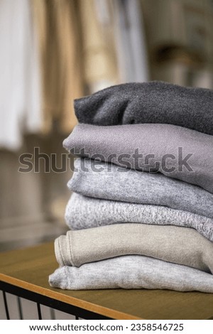 Stack of clean freshly laundered, neatly folded women's clothes on wooden table. Pile of shirts and sweaters on the table. Copy space, close up. Background rail with neutral clothes.
