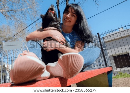 happy young black haired latin woman sitting on bench with her dog smiling and playing in park at sunset, enjoying day, copy space.