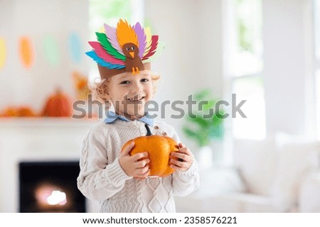 Child celebrating Thanksgiving. Kid holding pumpkin in paper turkey hat. Autumn fun crafts and art. Little blond curly boy in decorated living room. Warm knitted wear. Fall season decoration.