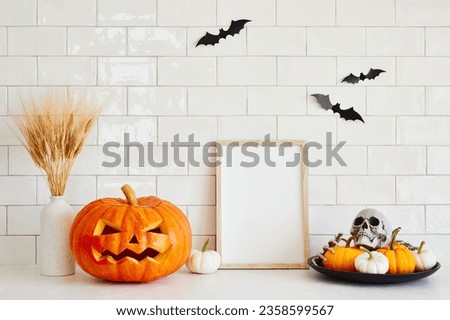 Halloween carved pumpkin, blank picture frame, vase of dried flowers, bats, decoration on white brick wall background. Halloween home decor.
