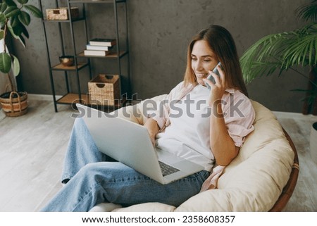 Young IT woman wear casual clothes sits in armchair hold use work on laptop pc computer talk on mobile cell phone stay home hotel flat rest spend free spare time in living room indoor. Lounge concept Royalty-Free Stock Photo #2358608357