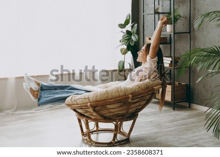 Full body side view young woman wear casual clothes sits in armchair raise up legs stretch hands stay at home hotel flat rest relax spend free spare time in living room indoor Lifestyle lounge concept Royalty-Free Stock Photo #2358608371