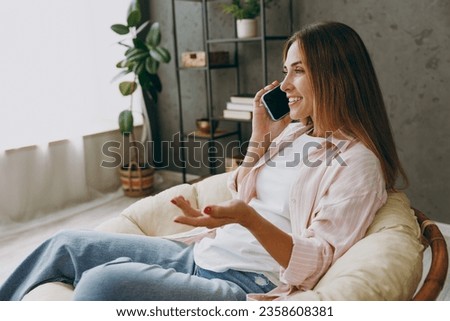 Side view young fun woman wear casual clothes talk speak on mobile cell phone sits in armchair stay at home hotel flat rest relax spend free spare time in living room indoor. Lifestyle lounge concept Royalty-Free Stock Photo #2358608381