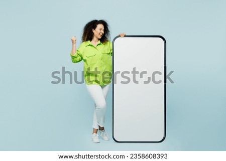 Full body young latin woman she wear green shirt casual clothes big huge blank screen mobile cell phone smartphone with workspace area do winner gesture isolated on plain pastel light blue background