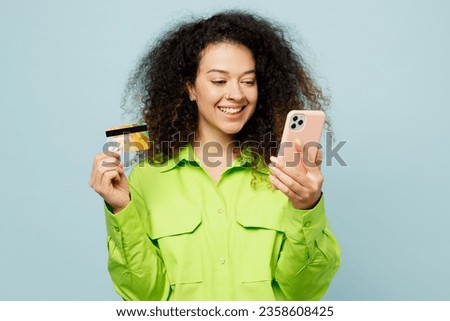 Young latin woman wear green shirt casual clothes using mobile cell phone hold credit bank card shopping online order delivery booking tour isolated on plain pastel light blue cyan background studio