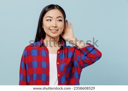 Young smiling curious nosy woman of Asian ethnicity 20s wear checkered shirt try to hear you overhear listening intently isolated on plain pastel light blue color background. People lifestyle concept