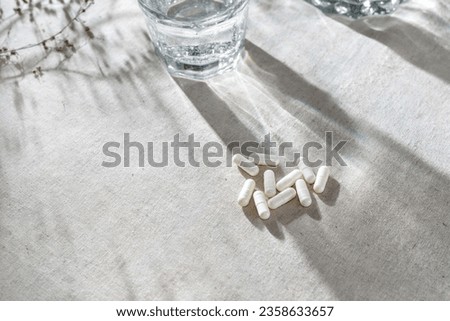 White capsules, glass with water and dried grass on neutral beige linen tablecloth with aesthetic light shadows. Royalty-Free Stock Photo #2358633657