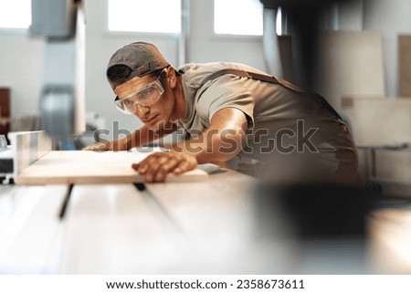Young carpenter cutting a piece of wood in using a circular saw in furniture factory Royalty-Free Stock Photo #2358673611