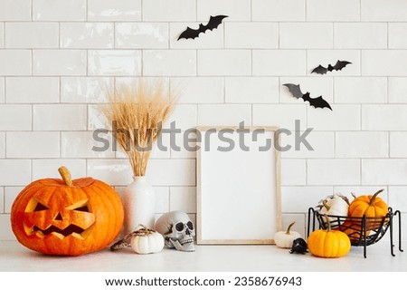 Halloween holiday home decor on white table. Picture frame mockup, vase of dried flowers, carved pumpkin, skull.