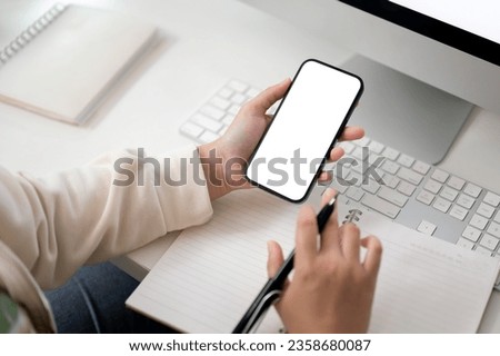 Close-up image of a female using her smartphone and taking notes on her notebook at her computer desk. smartphone white screen mockup