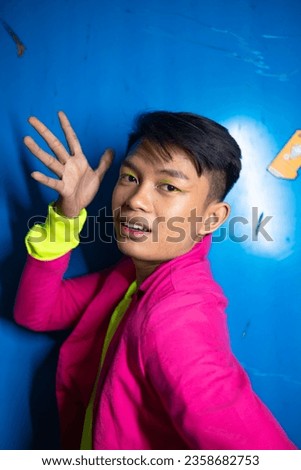 a gay Asian man posing very funny and humorous while wearing a pink suit in front of a blue background at night