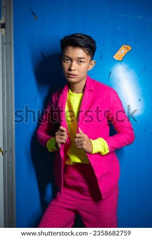 a gay Asian man posing very funny and humorous while wearing a pink suit in front of a blue background at night