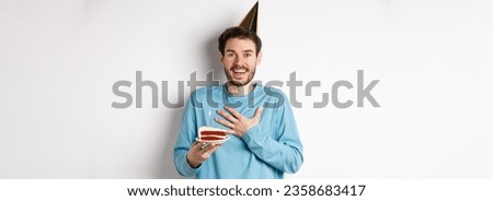 Celebration and holidays concept. Surprised birthday boy in party hat, holding bday cake and looking thankful, standing over white background.