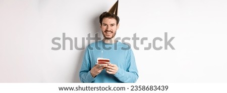Celebration and holidays concept. Happy young man in party hat celebrating birthday, holding bday cake and smiling, standing over white background.