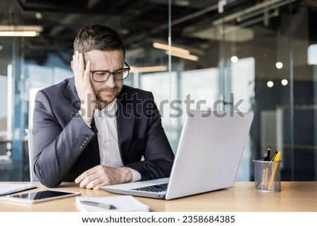 Problems at work. Upset young male office worker, businessman sitting at a desk in front of a laptop and thoughtfully leaning his head on his hand. Royalty-Free Stock Photo #2358684385