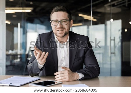 Portrait of a young businessman, a business coach conducts training and online training, professional development. Sitting in the office at the table in front of the camera, gesturing with his hands. Royalty-Free Stock Photo #2358684395
