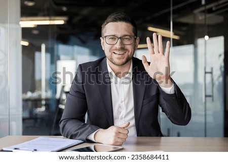 Portrait of a young businessman in a suit who sits in the office in front of the camera, communicates online, greets with his hand. Royalty-Free Stock Photo #2358684411