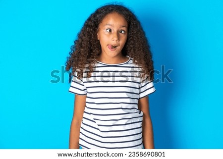 Funny beautiful kid girl with afro curly hairstyle wearing striped T-shirt makes grimace and crosses eyes plays fool has fun alone sticks out tongue.
