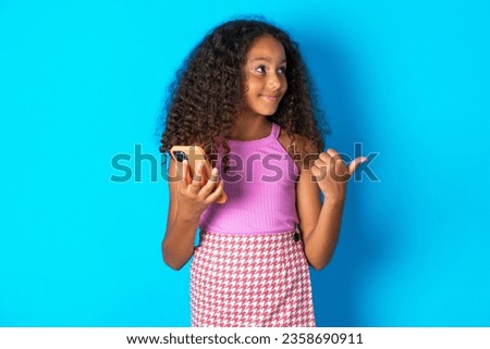 beautiful kid girl with afro curly hairstyle wearing pink T-shirt using and texting with smartphone  pointing and showing with thumb up to the side with happy face smiling
