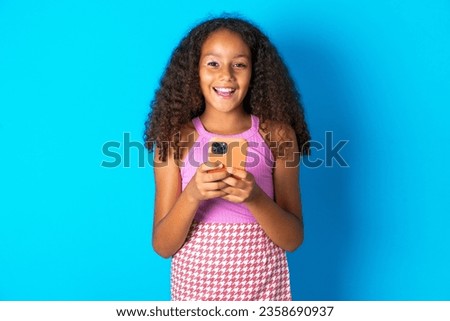 Excited beautiful kid girl with afro curly hairstyle wearing pink T-shirt holding smartphone and looking amazed to the camera after receiving good news.