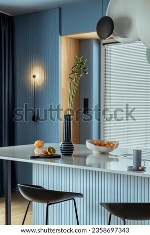 Interior design of kitchen interior with marble kitchen island, blue wall, black chokers, bowl with fruits, big window, cup, wooden floor, lamp on wall and personal accessories. Home decor. Template. Royalty-Free Stock Photo #2358697343