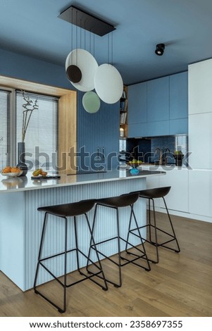 Interior design of kitchen interior with marble kitchen island, blue wall, black chokers, bowl with fruits, big window, cup, wooden floor, lamp on wall and personal accessories. Home decor. Template. Royalty-Free Stock Photo #2358697355