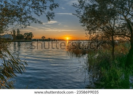 Sunset on the shore of Lake Constance in the Rhine Valley, Vorarlberg, Austria. sun is reflected in the water, framed by reeds and trees. Evening red, with red and orange clouds. recreation afternoon Royalty-Free Stock Photo #2358699035
