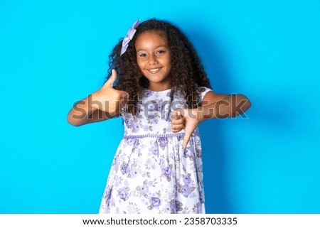 beautiful kid girl with afro curly hairstyle wearing floral dress showing thumbs up and thumbs down, difficult choose concept
