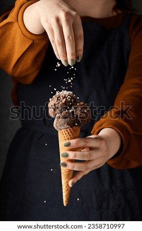 Woman sprinkling sea salt on top of dark chocolate ice cream. She is holding the ice cream cone with her other hand. Her torso is a background for the image. Dark lifestyle vertical picture.