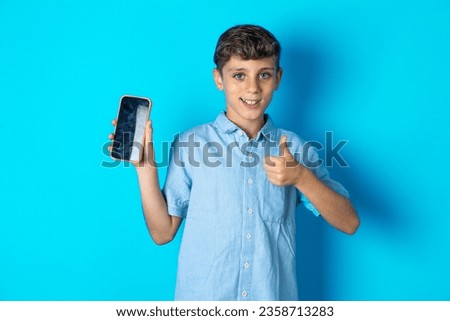 beautiful kid boy wearing casual clothes thinks deeply about something, uses modern mobile phone, tries to made up good message, keeps index finger near lips.