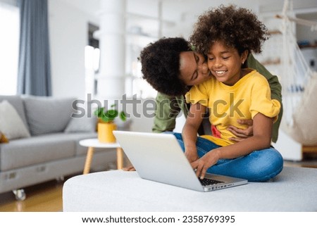 Loving African American mother kissing her young daughter who is using laptop at home. Royalty-Free Stock Photo #2358769395