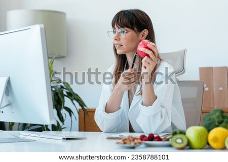 Shot of beautiful smart nutritionist woman doing videocall with computer to doing a pacient consultation in the nutritionist consultation Royalty-Free Stock Photo #2358788101