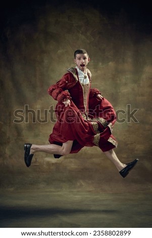 Dramatic apparel. Young man, royal person running on female dress on dark green background. Concept of historical retrospectives, fashion, provoking projects, gender fluidity, masculinity, femininity Royalty-Free Stock Photo #2358802899
