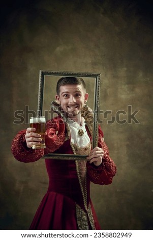 Medieval royal person, prince in female dress holding picture frame and drinking beer against dark green background. Concept of historical retrospectives, fashion, provoking projects, festival
