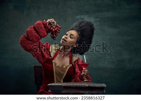 Winery. Young african woman, princess, queen in elegant red dress eating grapes and drinking wine against vintage green background. Concept of history, beauty and fashion, comparison of eras, ad