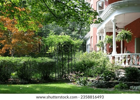 Victorian red brick house. Porch. Secret garden. Hanging baskets. Flowers. Vines. Trees. Wealthy. Historic. Hosta. Shrubs. Bushes. Black metal gate. Trees. Fall colour color. Leaves turning.  Royalty-Free Stock Photo #2358816493