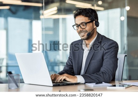 Young successful man at workplace with headset phone typing on laptop keyboard, customer support worker for online buyers inside office, hispanic helping and consulting customers. Royalty-Free Stock Photo #2358820147
