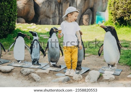 An adorable little boy holding icecream posing with little statues of penguins at a zoo