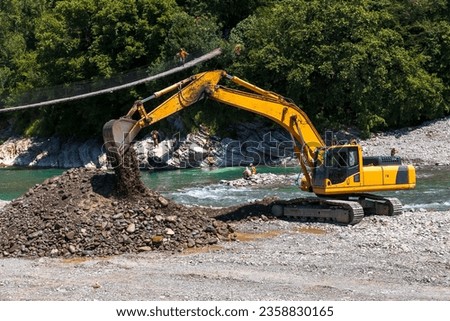 The excavator is working on expanding the riverbed. The work of heavy machinery. The use of heavy construction equipment. A yellow excavator at a construction site near the city river.  Royalty-Free Stock Photo #2358830165