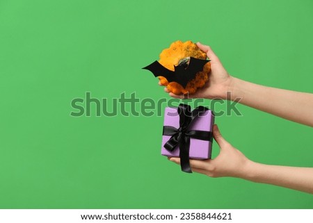 Female hands with gift box, Halloween pumpkin and paper bat on green background