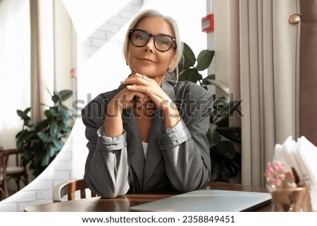 confident well-groomed senior woman with gray hair and glasses dressed in a gray jacket sits in a cafe at a table with a laptop Royalty-Free Stock Photo #2358849451