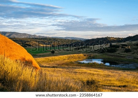 An early morning light glows over a pasture and hay farm with a pond in the front and mountains in the background.