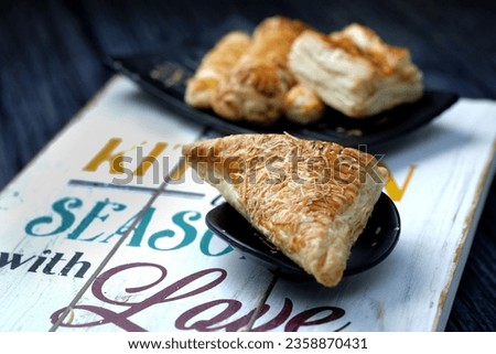 
A triangular shaped pastry filled with cheese and topped with cheese plating on a small plate sits on a wooden board with a poem about a kitchen with love, and in low light settings