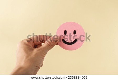A hand holding pink happy face in beige background with copy space. Positive customer feedback and review. Happiness and Good service concept.