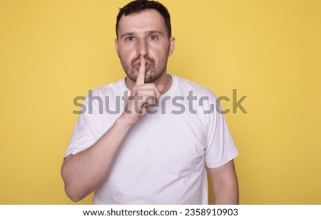 Young guy in white t-shirt hides secret and shows gesture of silence, man covers his mouth with his finger asks to be quieter on yellow isolated background. 