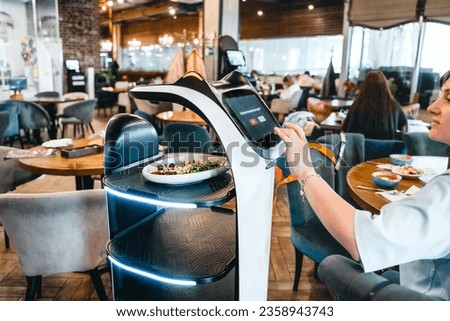 Robot waiter serve food at modern restaurant table.Offering innovation futuristic high-tech automated dining experience.Bringing,delivery automation order to customer.Digital robotic AI smart service. Royalty-Free Stock Photo #2358943743