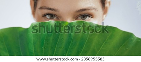 Natural skincare beauty portrait photo of a beautiful young woman hiding her face behind a green palm leaf while looking at the camera. High-quality adv studio photo of natural cosmetics and beauty Royalty-Free Stock Photo #2358955585