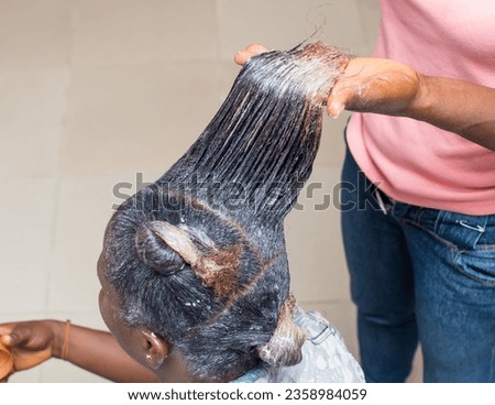 Hand of an African stylist, holding, displaying and applying relaxer cream to the natural long hair of a woman or female customer at a beauty salon in Nigeria Royalty-Free Stock Photo #2358984059