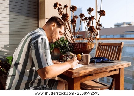 Side view of serious bearded male sitting at wooden table with mug of hot drink and yarn while taking notes